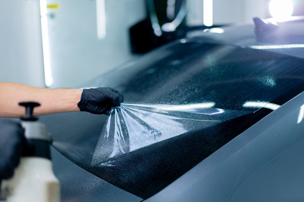 Film de protection PPF : Pose professionnelle sur voiture. Main gantée noire appliquant un film protecteur transparent (PPF) sur la lunette arrière humide d'une voiture sombre en atelier de detailing.