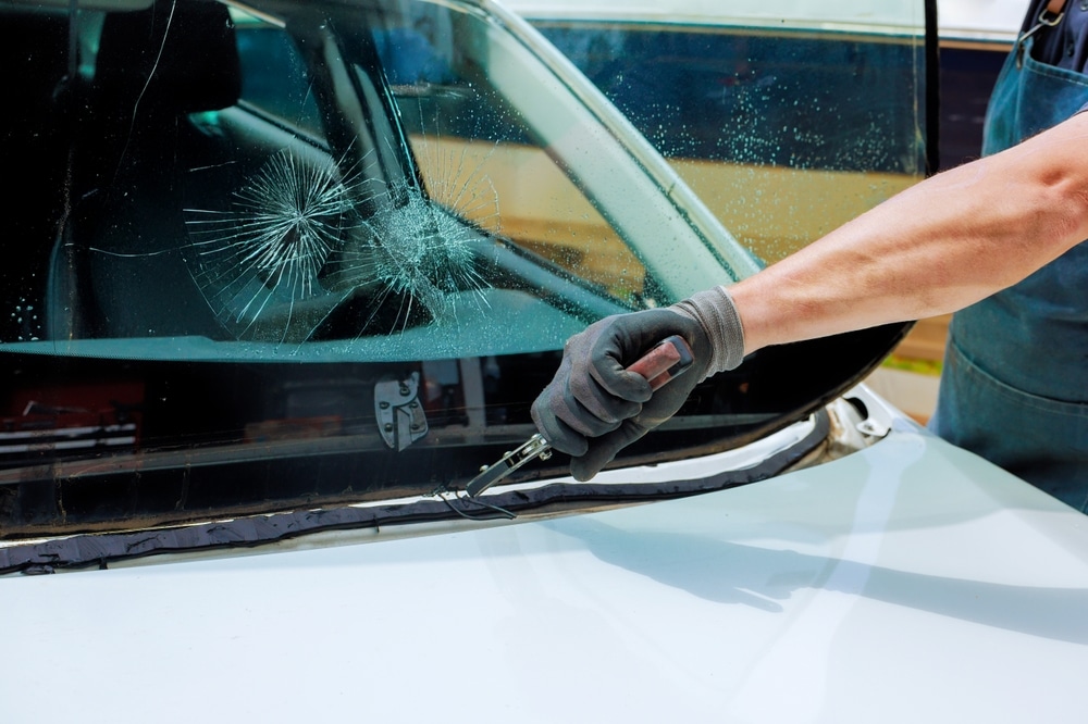 Pare-brise cassé : remplacement professionnel et rapide. Enlèvement d'un pare-brise de voiture blanc brisé (bris de glace) par un technicien coupant le joint d'étanchéité avec un outil.